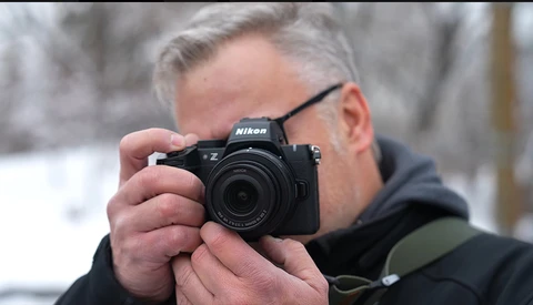 Photographer holding a Nikon Z mirrorless camera up to his face while wearing glasses and dark winter clothing.