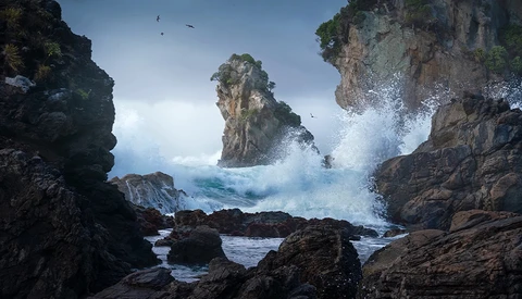 Dramatic ocean waves crashing against rocky sea stacks and cliffs under a partly cloudy sky.