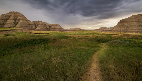 Dirt trail cutting through grassland with layered buttes under dramatic storm clouds.
