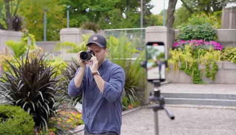 Photographer holding camera to face while looking through viewfinder in manicured garden setting.