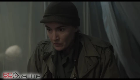 Person wearing dark fedora hat and olive jacket in dimly lit interior space.