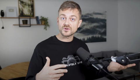 Man in black camera-themed t-shirt speaking expressively while holding a microphone in an indoor studio setting.