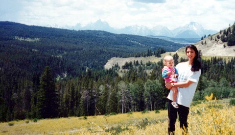 Woman holding a toddler standing in a mountain meadow with forested valleys and snow-capped peaks in the background.