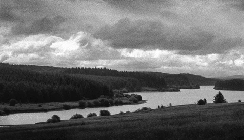 Dramatic landscape with forested hills, a calm lake, and storm clouds overhead in black and white.