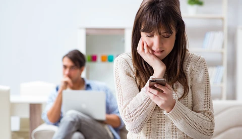 Woman in cream cardigan smiling while looking at smartphone, with man blurred in background working on laptop.