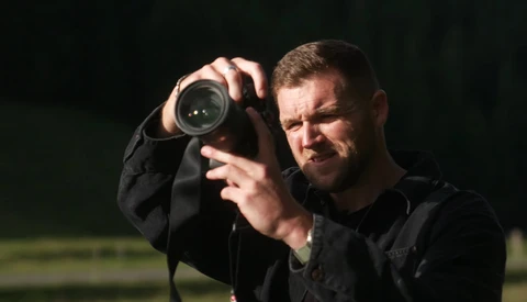 Photographer holding binoculars to his face against a dark background.