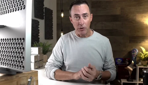 Man in light gray long-sleeve shirt speaking to camera in modern home studio setting.