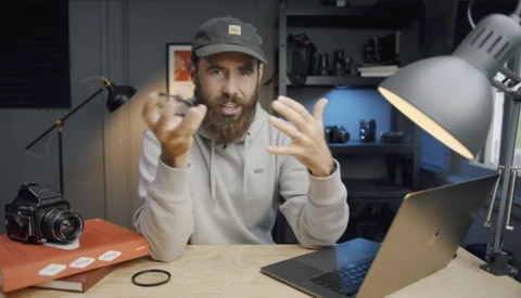 Bearded man in cap gesturing while sitting at desk with camera, laptop, and lighting equipment.