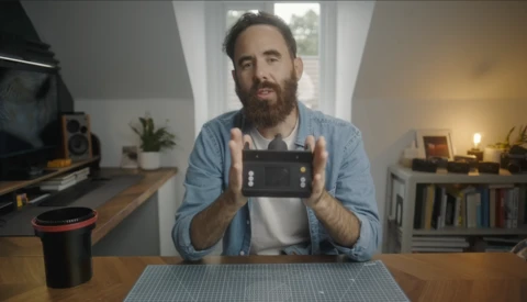 Bearded man holding a camera controller while seated at a desk in a home studio.