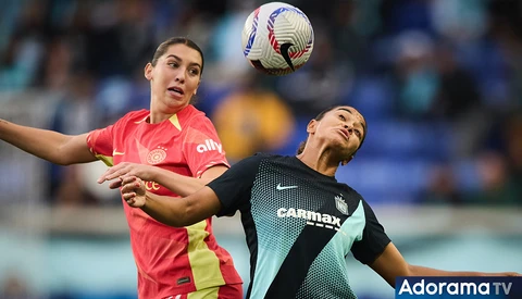 Two female soccer players competing for a header, one in red and yellow jersey, one in black and teal jersey.