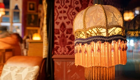 Ornate vintage pendant lamp with cream fabric shade, gold embroidered trim, and long orange beaded fringe hanging in warm interior space.