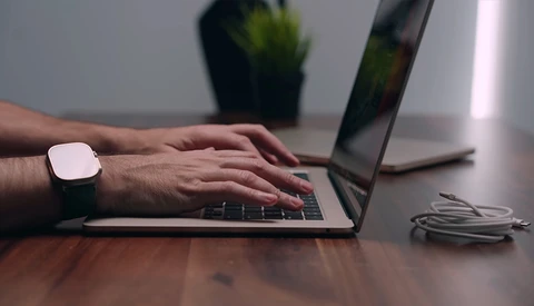 Person typing on laptop keyboard while wearing a smartwatch at a wooden desk.
