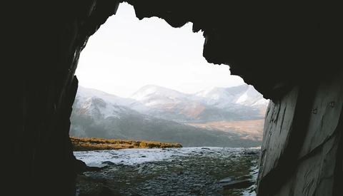 Snow-capped mountains and a rocky coastline viewed through a natural cave opening.