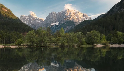Alpine lake with forested shoreline and snow-capped mountain peaks reflected in still water.