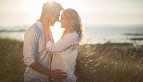 Couple embracing during golden hour on a beach with tall grass and ocean backdrop.