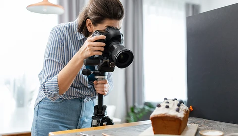 Female photographer holding a mirrorless camera with telephoto lens mounted on a tripod, shooting a styled food setup.