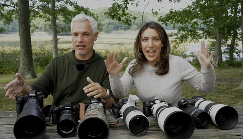 Two people display a collection of camera lenses arranged on a wooden table outdoors.