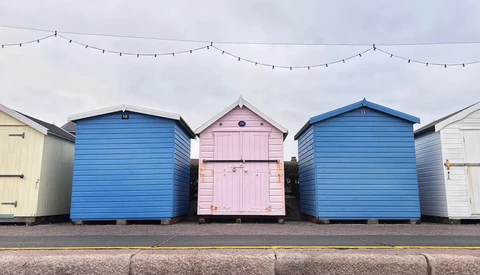 Three colorful beach huts in a row with blue and pink painted wooden facades and peaked roofs.
