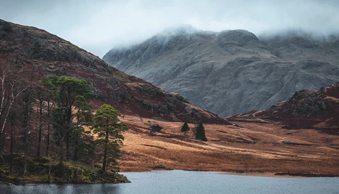 Misty mountains rise above a moorland valley with a small loch in the foreground and scattered trees along the shoreline.