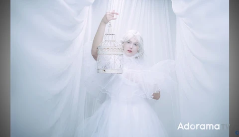 Woman in white ethereal dress holding ornate birdcage amid flowing white fabric and soft lighting.