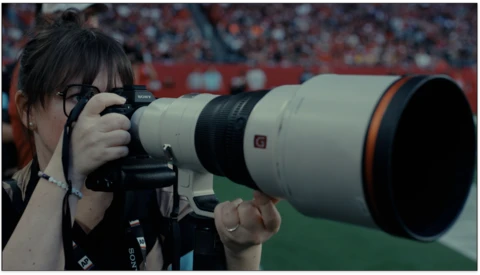 Sports photographer with telephoto lens at a stadium event with crowd in background.