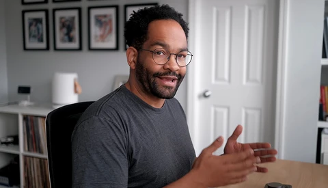 Man wearing glasses and dark t-shirt gesturing while speaking in a home office setting.