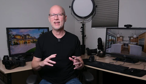 Bald man with glasses speaking to camera in a photography studio setup with dual monitors and professional lighting equipment.