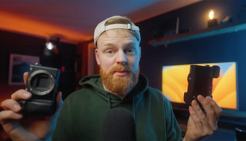 Photographer holding a camera body in one hand and a smartphone in the other, displayed in a studio with warm and cool ambient lighting.