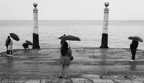 Five people with umbrellas stand on a wet promenade near decorative pillars overlooking calm water.