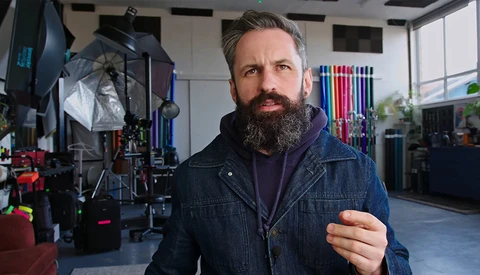 Bearded man in denim jacket inside a photography studio with lighting equipment and reflectors visible behind him.