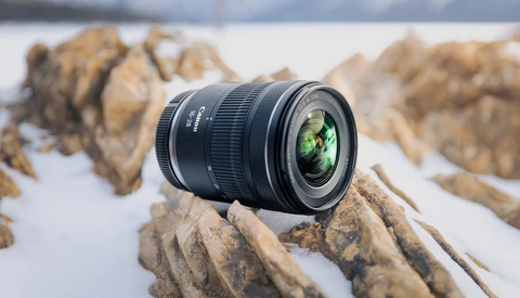 Wide-angle zoom lens positioned on rocky terrain with snowy background and blurred mountains.