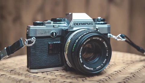 Olympus OM10 35mm film camera with black leather grip and attached Zuiko lens on woven surface.