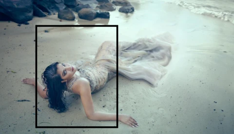 Woman in flowing white dress lying on wet sand at shoreline with waves and rocks visible.