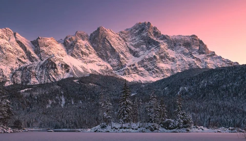 Snow-covered alpine peaks glowing pink at sunset above a frozen lake and evergreen forest.