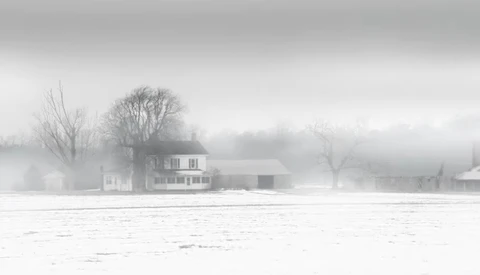 Farmhouse in heavy fog with bare trees and snow-covered field in winter.