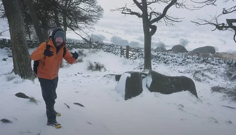 Runner in orange jacket jogging through snowy rural landscape with bare trees and stone walls.