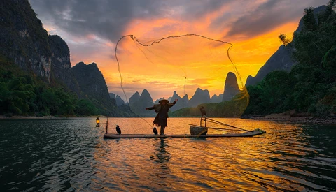 Fisherman casting net at sunrise among dramatic limestone karst formations reflected in still water.