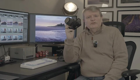 Photographer in studio holding DSLR camera with studio lighting setup visible overhead.