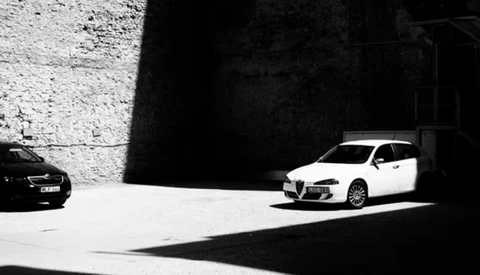 White sedan parked in stark sunlight against concrete wall with deep shadow in monochrome.