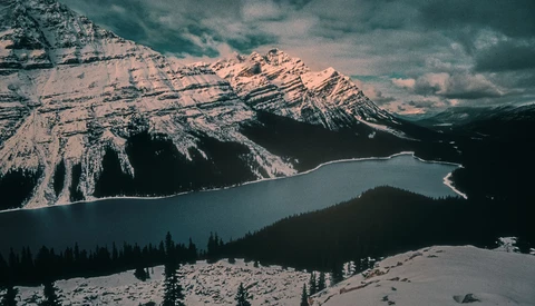 Snow-covered mountain peaks tower above a turquoise alpine lake surrounded by evergreen forest.