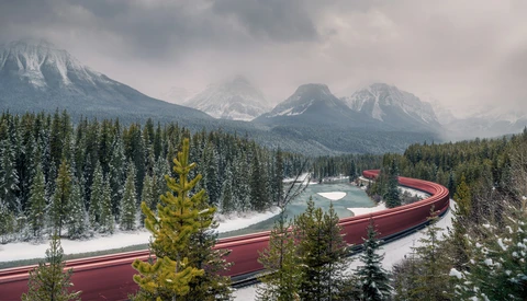 Snow-covered mountain landscape with coniferous forest and red train curving through valley.