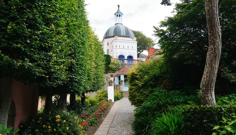 Manicured garden pathway lined with green hedges leading to a domed building with classical architecture.