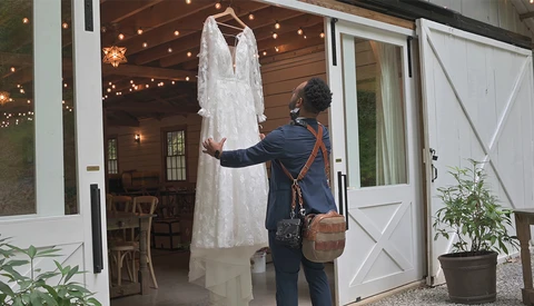 Woman in dark blazer examining a white lace wedding dress hanging indoors.