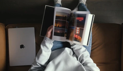 Overhead view of a person in a white hoodie holding an open photography magazine or portfolio while sitting on a couch with an iPad nearby.