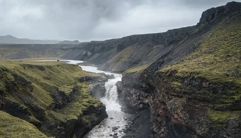 Glacial river cutting through moss-covered canyon with basalt cliffs under overcast sky.
