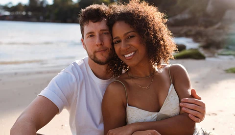 Couple embracing on a sandy beach during golden hour with water and shoreline in background.