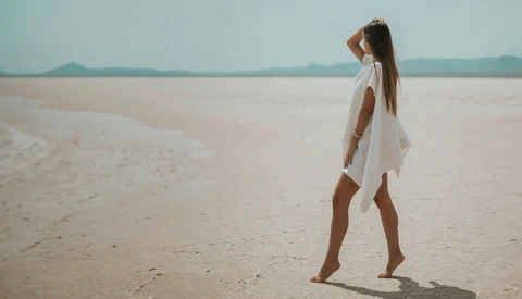 Woman in white dress standing barefoot on expansive sandy beach, gazing toward distant horizon and mountains.