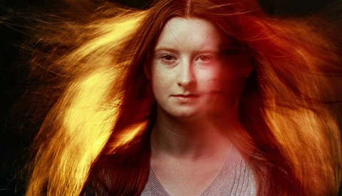 Portrait of a woman with long red-orange hair against a dark background with warm golden lighting.