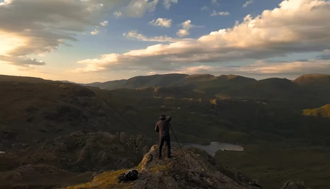 Photographer on rocky outcrop with tripod overlooking mountainous valley at golden hour.