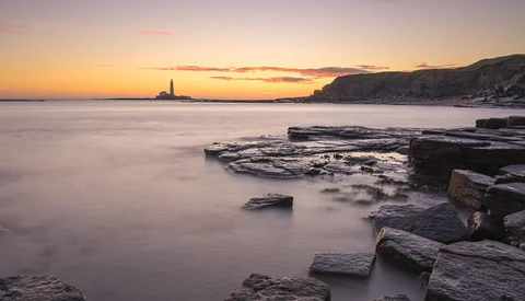 Rocky coastline at sunset with calm water and lighthouse in the distance.